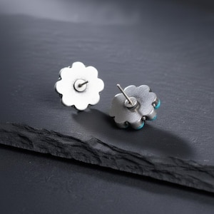 May include: A pair of silver-coloured flower stud earrings with turquoise accents. The earrings have a detailed, scalloped edge design. The posts are visible on the back of the earrings. The earrings are displayed on a dark, textured surface.