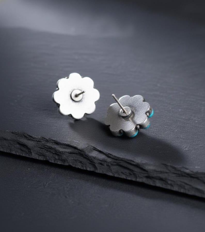 May include: A pair of silver-coloured flower stud earrings with turquoise accents. The earrings have a detailed, scalloped edge design. The posts are visible on the back of the earrings. The earrings are displayed on a dark, textured surface.