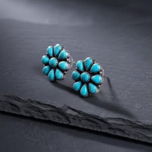 May include: A pair of silver stud earrings with a floral design. Each earring has a cluster of turquoise-coloured, teardrop-shaped stones. The earrings are set against a dark background, highlighting the bright blue stones.