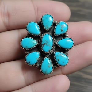 May include: A silver-toned ring featuring a cluster of turquoise stones. The ring's design showcases a central oval stone surrounded by eight teardrop-shaped stones, all set in a detailed metalwork setting. The turquoise stones have a vibrant blue-green hue.