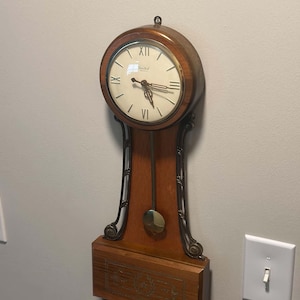 May include: A wall-mounted wooden clock with a circular face featuring Roman numerals and the text United. The clock has a brass-colored pendulum, ornate metal side supports, and a rectangular base decorated with a floral inlay pattern.