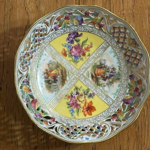 May include: An antique, decorative porcelain plate with a scalloped edge and intricate openwork design. The plate features a yellow center with floral and pastoral scenes, surrounded by colorful flowers and a gold trim.