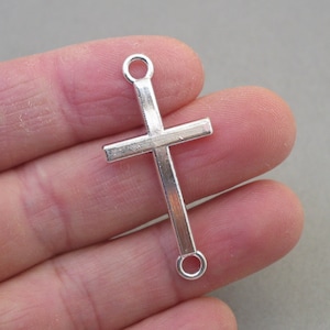 May include: Silver-toned metal cross charm with two jump rings. The cross is flat and has a simple design.