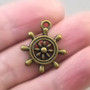 May include: A small, bronze-colored ship's wheel charm. The charm has eight spokes and a central floral design. It is a nautical-themed accessory, perfect for jewelry making or crafting projects. The charm is held in a person's hand.