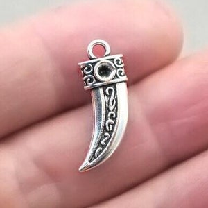 May include: A silver-colored pendant in the shape of a curved tooth. The top of the pendant has a decorative band with a circular design and a small loop for attaching to a chain. The tooth has etched details.