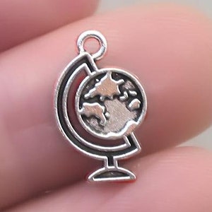 May include: A small, silver-colored globe charm with a detailed world map design. The charm is attached to a small loop at the top, and the globe is held by a stand. The charm is held between two fingers.