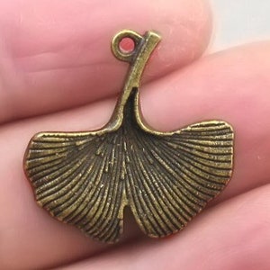 May include: A bronze-colored metal charm in the shape of a ginkgo leaf. The charm has a small loop at the top for attaching to a chain or other jewelry. The leaf has detailed veining and a slightly aged appearance.