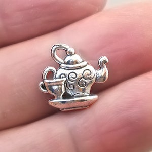 May include: Silver charm in the shape of a teapot and cup. The teapot has a lid and a spout. The cup is on a saucer. The charm is detailed with swirls and lines.