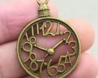 May include: A bronze-colored metal clock face charm with Roman numeral markings and a cutout design. The charm has a loop at the top for attaching to a necklace or bracelet.