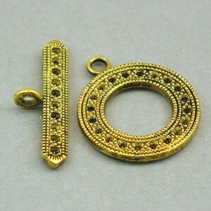 Peut inclure: Un ensemble de fermoir à bascule de couleur dorée. Le fermoir comprend une barre rectangulaire avec une série de petits trous et un anneau circulaire avec un motif complexe. La trouvaille de bijoux est conçue pour connecter deux pièces.