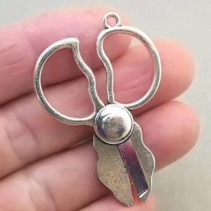 May include: Silver-toned metal pendant shaped like a pair of scissors with a loop at the top for attaching to a chain or cord.