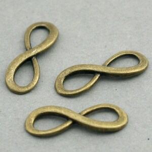 May include: Three bronze-coloured infinity symbol charms. Each charm has a smooth, rounded design. The charms are arranged on a light grey surface, with varying orientations. The metal has a slightly aged appearance.