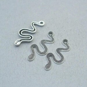 May include: Silver-toned snake charms in various sizes. One charm has a black outline, while the others have a textured, dotted pattern. These charms are ideal for jewelry making or crafting projects.