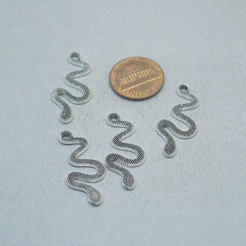 May include: Five silver-colored snake charms with a textured, scaled design. A penny is included for size reference. The charms are small and have a loop at the top for attaching to jewelry or other crafts.