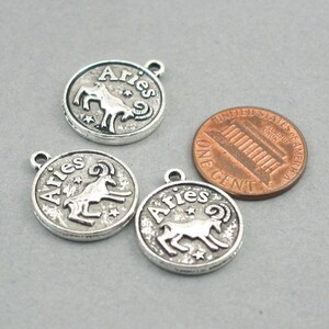 May include: Three silver-toned metal charms with the zodiac sign Aries and the word "Aries" written on them. The charms are round and have a textured surface. The charms are about the size of a US penny.