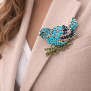 May include: A decorative bird brooch, featuring turquoise, blue, and clear crystals, set on a green branch. The brooch is attached to a beige blazer. The bird's design is eye-catching, and the brooch is a stylish accessory.