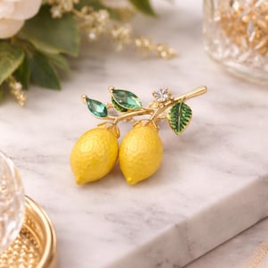 May include: A gold-toned brooch featuring two yellow lemon charms with green leaves and emerald-colored accents. The brooch is designed to resemble a lemon branch, perfect for adding a touch of nature-inspired elegance to any outfit.