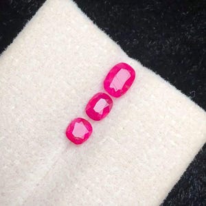 Può includere: Tre gemme rosse vivaci, taglio ovale, sono esposte su un tessuto bianco strutturato. Le gemme sono disposte in diagonale, mostrando le loro superfici lucide e la chiarezza interna. Lo sfondo è nero a contrasto.