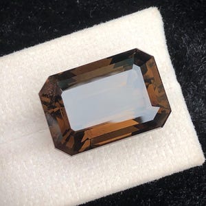 Puede incluir: Una gema rectangular facetada con un tono marrón ahumado. La gema está colocada sobre una tela blanca, con un fondo oscuro y texturizado. El corte de la piedra crea múltiples superficies reflectantes.