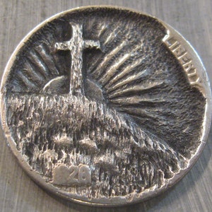 May include: A silver US penny with a cross on a hill and a sunburst behind it. The coin has been altered with a design that is raised from the surface. The coin has the word "LIBERTY" on the right side and the number "26" on the bottom.