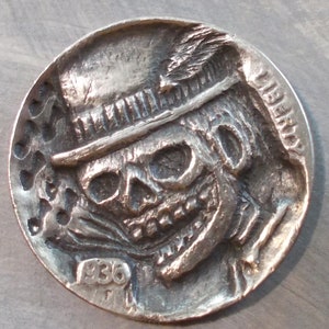 Könnte beinhalten: Eine silberfarbene Münze mit einem Schädel, der einen Zylinder trägt, der in die Oberfläche geschnitzt ist. Die Münze trägt die Aufschrift "LIBERTY" auf der Seite und das Jahr "1936" auf der Unterseite.