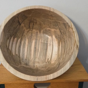 May include: A large, round wooden bowl with a natural wood grain pattern. The bowl is light brown with darker streaks and sits on a small wooden table. The bowl's interior is smooth and polished.