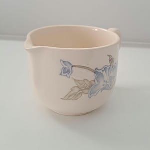 May include: Cream-colored ceramic creamer with a blue floral design. The creamer has a small handle and a spout for easy pouring. The floral pattern includes blue flowers and green leaves.