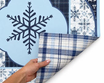 May include: A rectangular placemat featuring a large dark blue snowflake on a light blue background, surrounded by panels of dark blue plaid and white snowflake patterns. The corner is folded to reveal a light blue, white, and dark blue plaid underside.