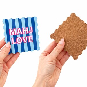 May include: A square coaster with scalloped edges featuring vertical light and dark blue stripes and the text MAHJ LOVE in pink letters. The reverse side of the coaster is made of brown cork with a matching scalloped edge design.