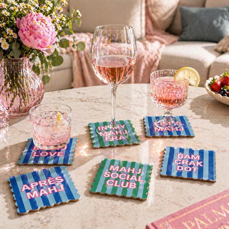 May include: Five square coasters with scalloped edges featuring blue and green vertical stripes and pink text. The text reads: IN MY MAHJ ERA, LET'S MAHJ, MAHJ LOVE, APR&Egrave;S MAHJ, MAHJ SOCIAL CLUB, and BAM CRAK DOT. The coasters are arranged on a table.