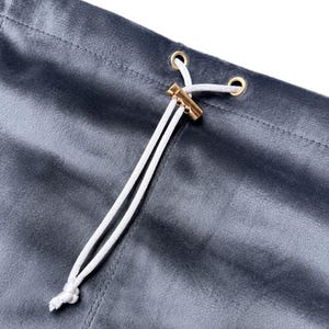 May include: A dark gray velvet fabric garment featuring a white drawstring cord threaded through two gold-toned metal eyelets. The cord is secured by a cylindrical gold-toned metal toggle, with the ends of the cord knotted at the bottom.