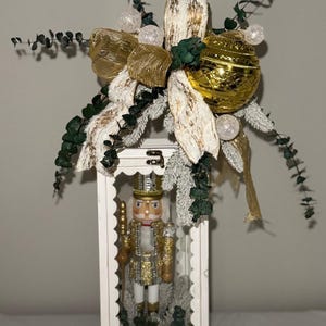 May include: A white wooden lantern with a gold and white Christmas decoration on top. Inside the lantern is a nutcracker soldier. The decoration includes gold ribbon, a gold ball, and sprigs of greenery. The lantern is a festive holiday decoration.