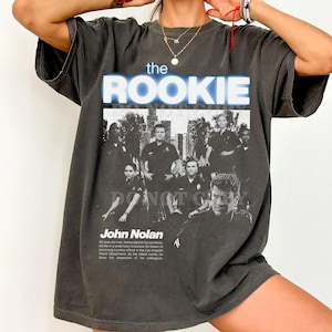 Może przedstawiać: Ciemnoszara koszulka z napisem "the ROOKIE" w niebieskim kolorze, z grupą osób w mundurach policyjnych. Poniżej tekstu znajduje się imię "John Nolan" i krótki opis.