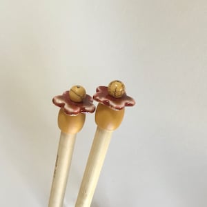 May include: Two wooden knitting needles with decorative flower-shaped tops. The tops are made of ceramic and have a brown and white glaze. The needles are natural wood color.