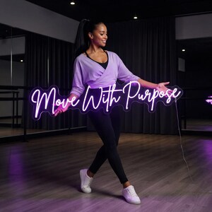 May include: A bright neon sign featuring the text Move With Purpose in a cursive white font. The sign is rectangular with a clear acrylic backing and a thin power cord extending from the right side, emitting a soft glow against the dark room.