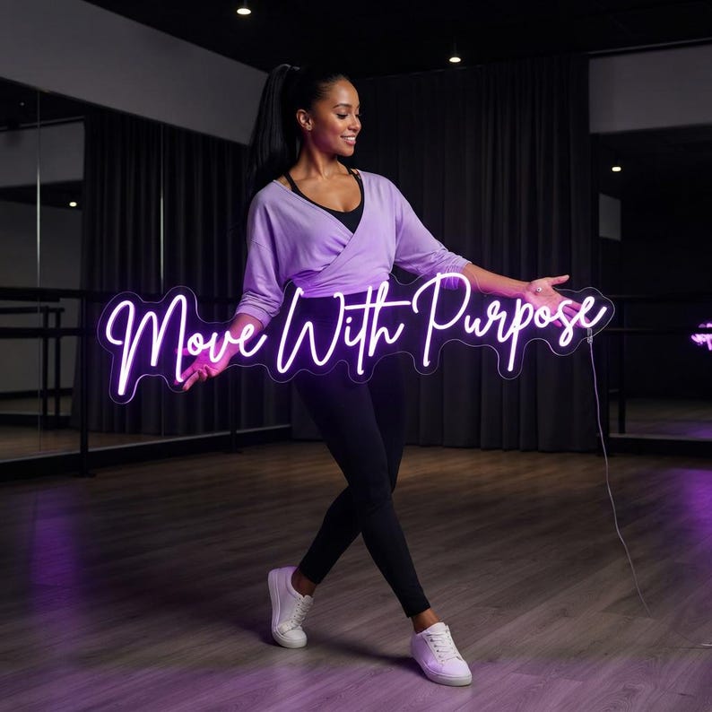 May include: A bright neon sign featuring the text Move With Purpose in a cursive white font. The sign is rectangular with a clear acrylic backing and a thin power cord extending from the right side, emitting a soft glow against the dark room.