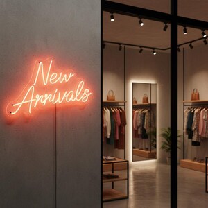 May include: A neon sign with the words "New Arrivals" in a cursive font, glowing orange against a gray wall. The sign is in a retail store, with clothing racks and a mirror visible through a glass partition.