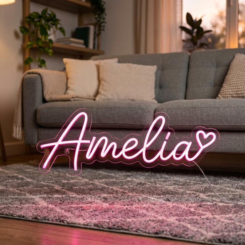 May include: A pink neon sign featuring the name Amelia in a cursive script font with a small heart symbol at the end. The sign is constructed from clear acrylic backing and illuminated pink tubing, connected to a thin power cord extending outward.