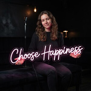 May include: A neon sign featuring the words Choose Happiness in a cursive script font. The sign emits a bright white glow and is mounted on a clear acrylic backing that follows the contour of the lettering. The text is clearly legible and centered.