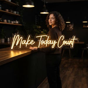 May include: A horizontal neon sign featuring the words "Make Today Count" in a warm, glowing yellow script font. The sign is mounted on a clear acrylic backing and emits a bright, steady light, displaying the text clearly against the dark background.