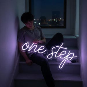 May include: A custom neon sign featuring the words one step in a flowing, cursive script font. The sign emits a bright, cool-toned white light that illuminates the surrounding area with a soft purple glow, mounted on a clear acrylic backing.