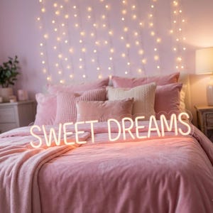 May include: A pink bed with a plush comforter and pillows. A neon sign that reads "SWEET DREAMS" is placed on the bed. String lights hang on the wall behind the bed, creating a cozy atmosphere.