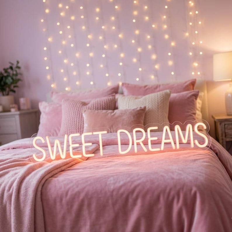 May include: A pink bed with a plush comforter and pillows. A neon sign that reads "SWEET DREAMS" is placed on the bed. String lights hang on the wall behind the bed, creating a cozy atmosphere.