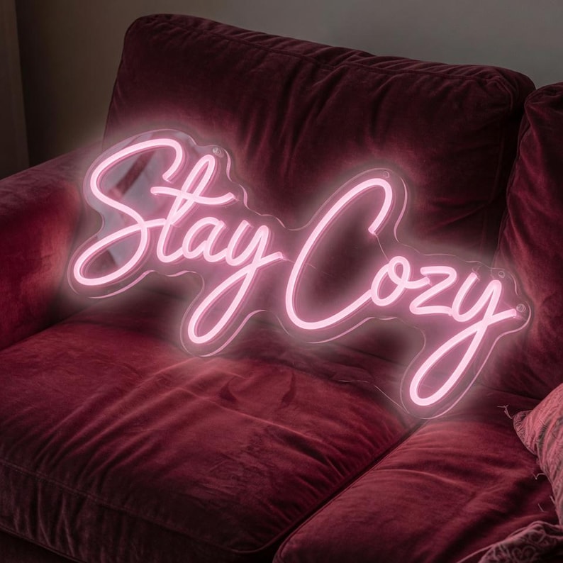 May include: A pink neon sign that reads "Stay Cozy" in a cursive font. The sign is illuminated and set against a dark red velvet couch. The sign is a decorative item for a home.