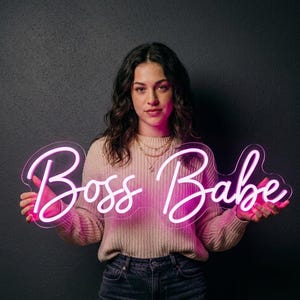 May include: A neon sign in a cursive font that reads "Boss Babe" in bright pink. The sign is held up against a dark background. The sign is lit up and the letters are easy to read.