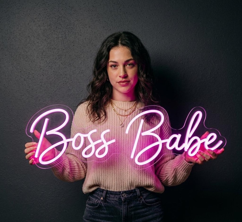 May include: A neon sign in a cursive font that reads "Boss Babe" in bright pink. The sign is held up against a dark background. The sign is lit up and the letters are easy to read.