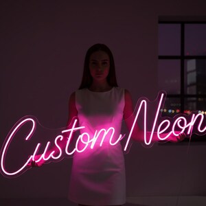 May include: A bright pink neon sign featuring the text Custom Neon in a cursive script font. The sign is mounted on a clear acrylic backing and emits a vibrant glow, illuminating the surrounding area with a distinct pink light.