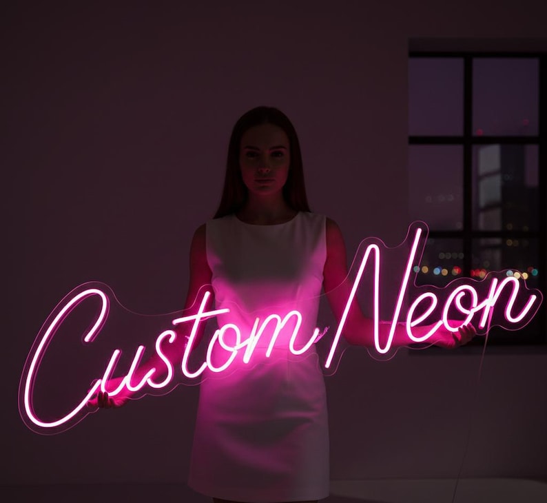 May include: A bright pink neon sign featuring the text Custom Neon in a cursive script font. The sign is mounted on a clear acrylic backing and emits a vibrant glow, illuminating the surrounding area with a distinct pink light.
