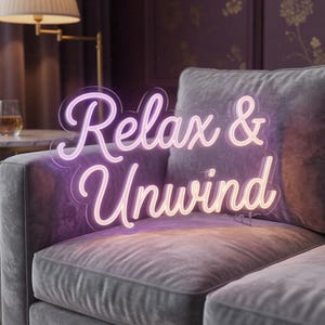 May include: A neon sign with the words "Relax & Unwind" in a cursive font, illuminated with a gradient of pink and white light. The sign is placed on a gray velvet sofa, creating a cozy and inviting atmosphere.