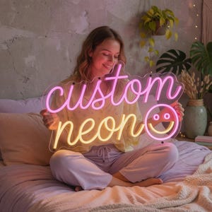 May include: A custom neon sign featuring the word custom in pink script, the word neon in yellow script, and a yellow smiley face icon. The sign is mounted on a clear acrylic backing and emits a bright glow, displaying the text custom neon.